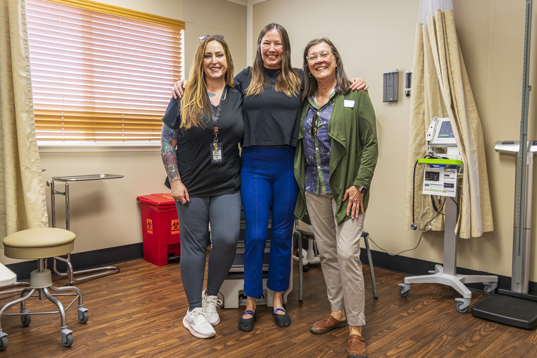 Three women stand together in a Panther Clinic exam room, with medical equipment and privacy curtains nearby.